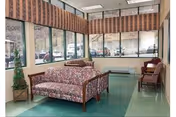 A waiting area with floral patterned sofas and chairs arranged near large windows with vertical blinds. There is a small potted plant in the corner and a green tiled floor.