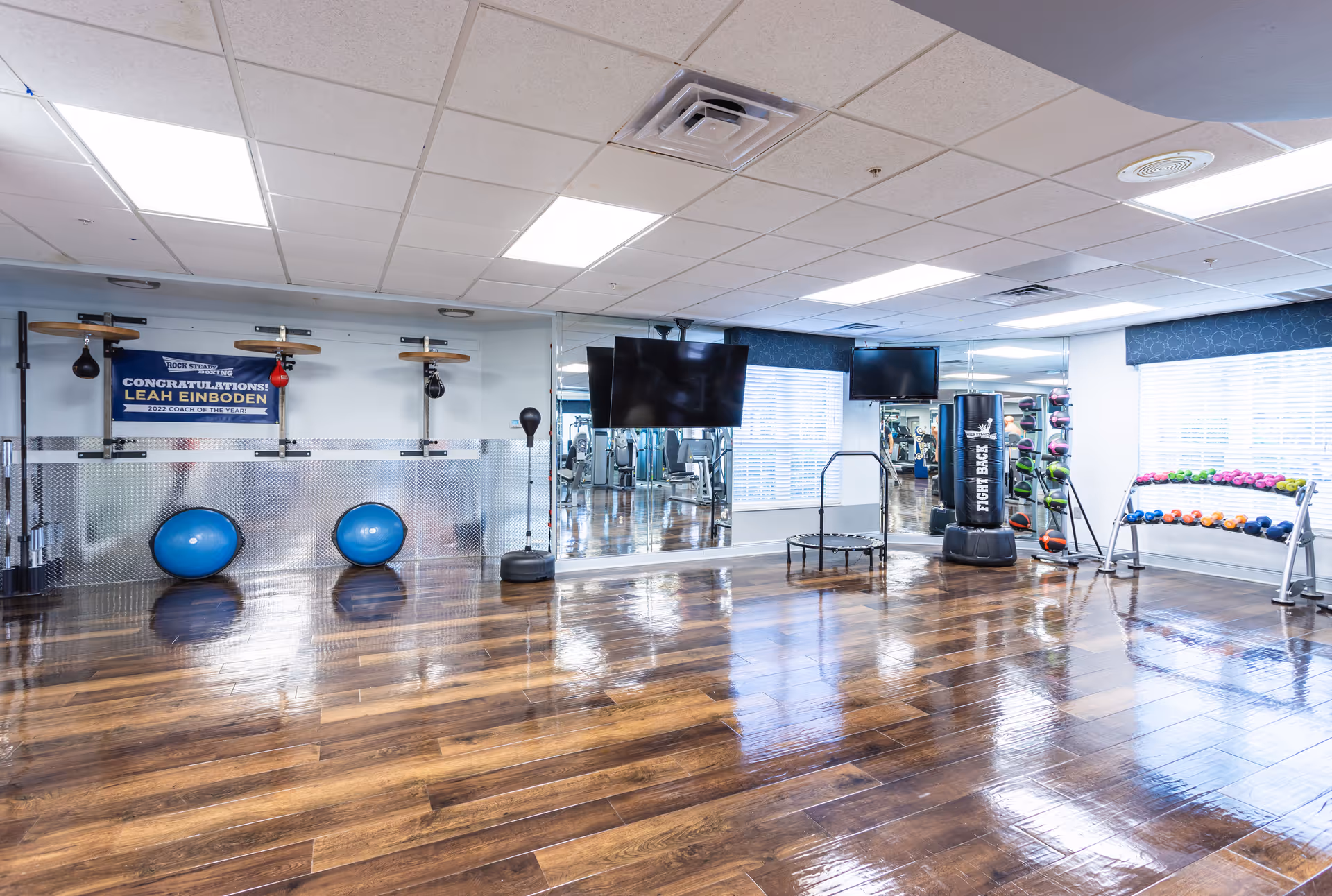 A spacious fitness room with polished wooden floors, two blue exercise balls, a punching bag, a small trampoline, and racks of colorful dumbbells and medicine balls. The room has large windows with blinds, two wall-mounted flat-screen TVs, and mirrors along one wall.
