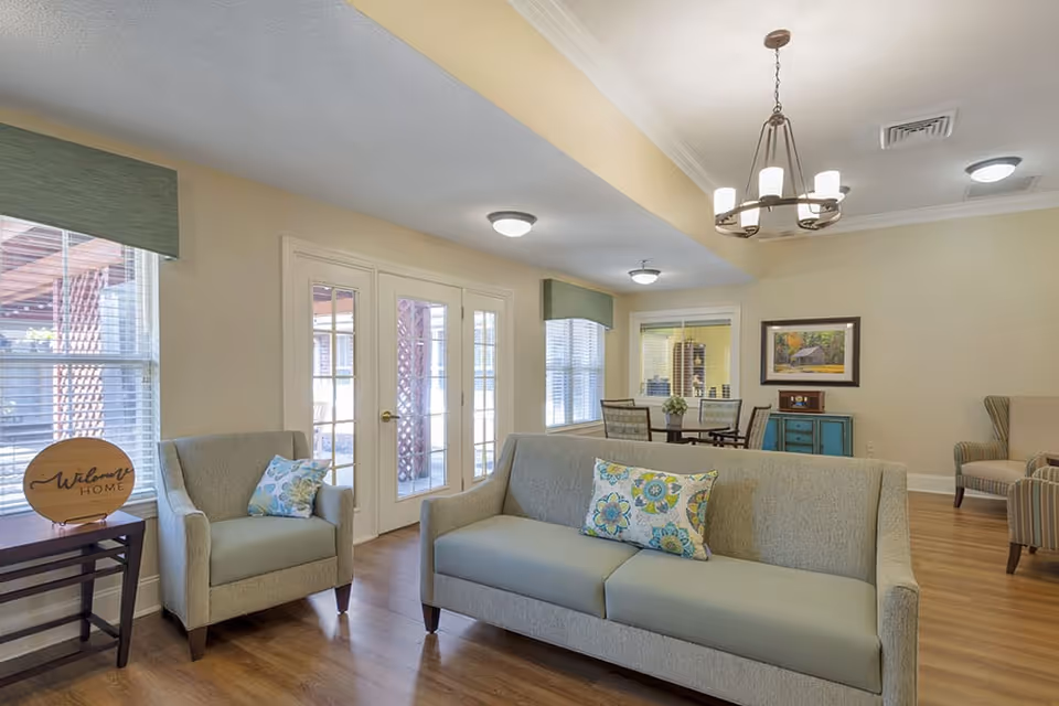A bright and welcoming living room area with a light gray sofa and matching armchair, both adorned with floral patterned cushions. There is a small wooden side table with a round wooden sign that says 'Welcome Home'. The room has wooden flooring, beige walls, and large windows with green valances. In the background, there is a dining table with four chairs, a blue sideboard with a framed picture above it, and additional seating. French doors lead to an outdoor area with a pergola.