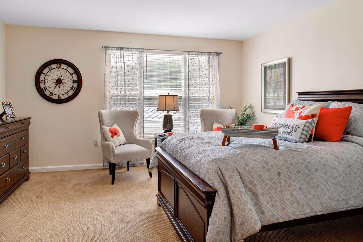A cozy bedroom with a large wooden bed adorned with gray and red pillows, including one with the text 'happiness is homemade.' There are two beige armchairs with red flower pillows near a window with sheer curtains. A wooden dresser with a framed photo and a large decorative wall clock are on the left side of the room. A bedside table with a lamp is positioned between the chairs.