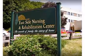 A green and gold sign reading "East Side Nursing & Rehabilitation Center" stands in front of shrubs with a multi-story building visible behind it.