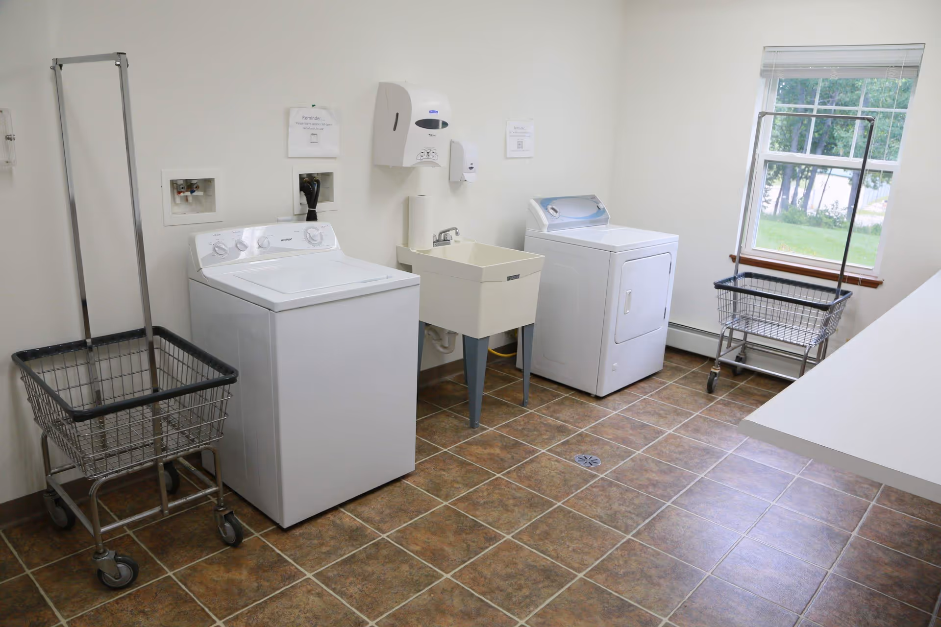 A clean laundry room with a washer, dryer, utility sink, two wheeled laundry carts, and a folding counter by a window.