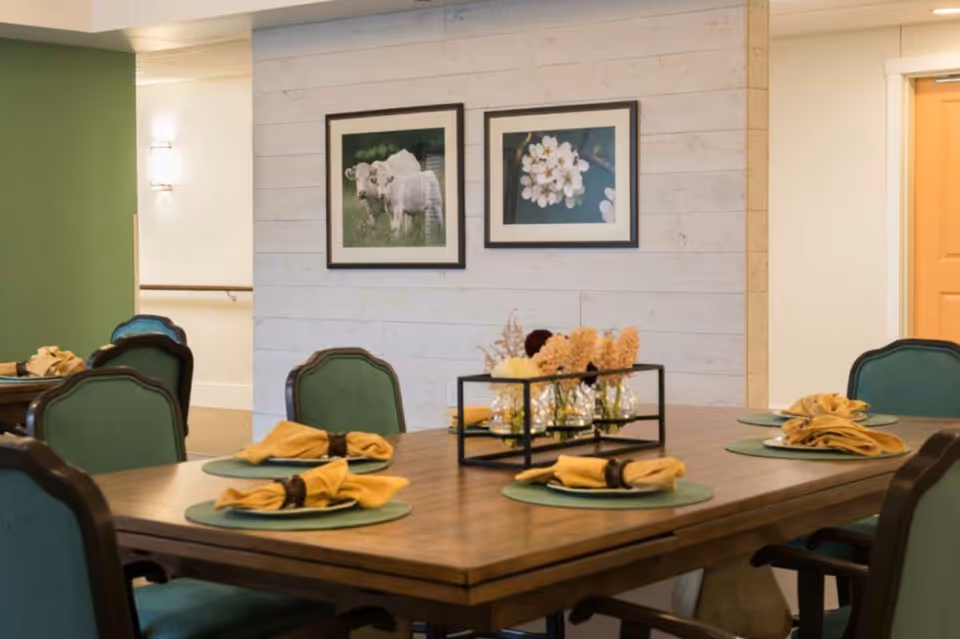 A dining room with a wooden table set for six, featuring green cushioned chairs, green placemats, and yellow napkins with napkin rings. A centerpiece with dried flowers in glass vases is on the table. The background shows a white shiplap wall with two framed pictures, one of cows and one of white flowers.