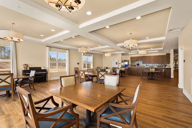 Spacious communal dining area with wooden tables and chairs, chandeliers, large windows, and a service counter in the background.
