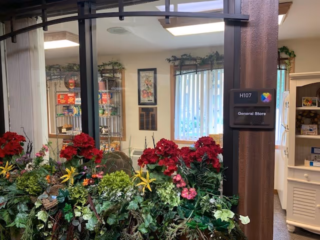 View through a glass window into a general store room labeled H107, with shelves stocked with various items and a variety of artificial flowers and plants arranged in front of the window.