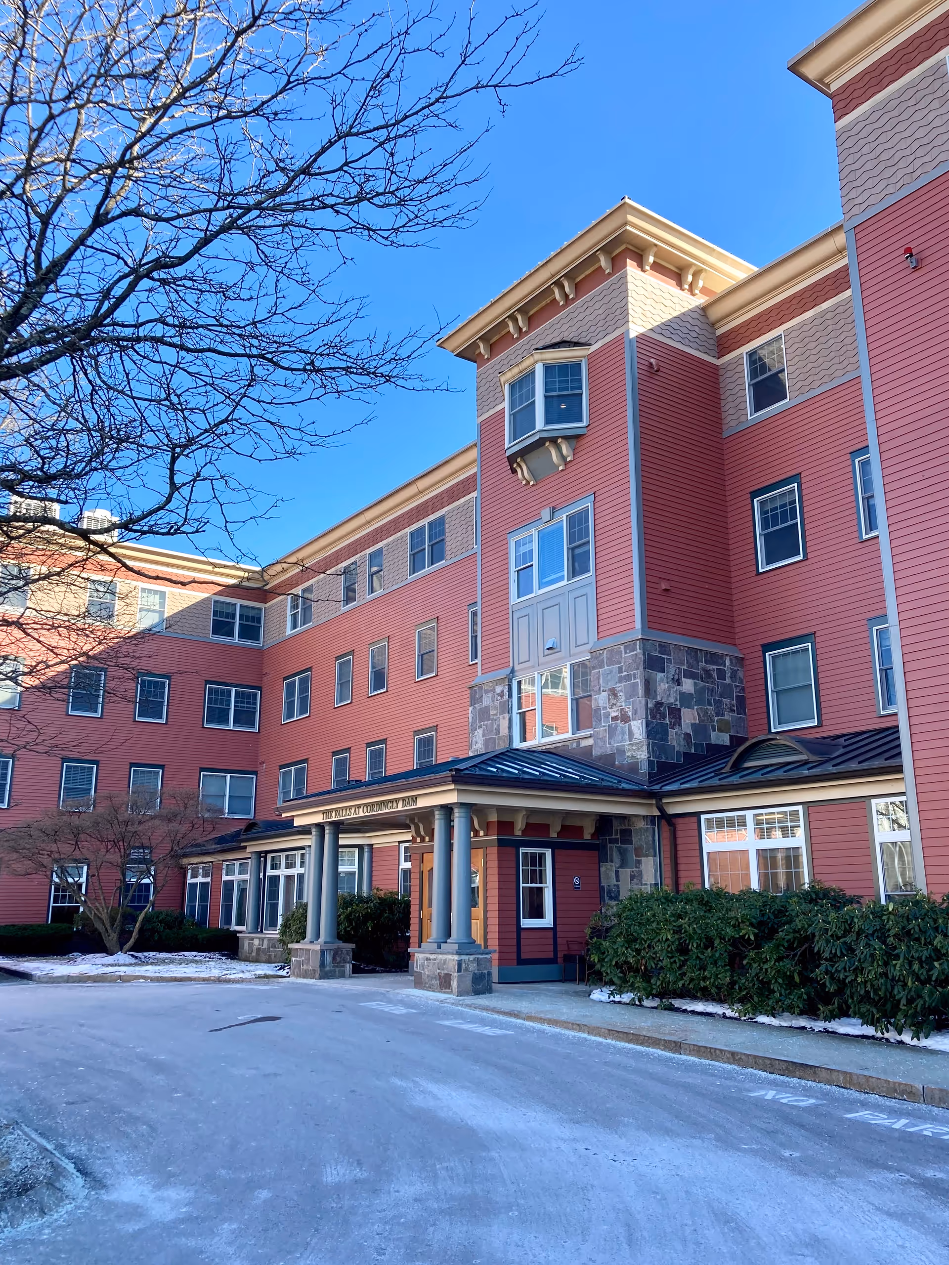 Exterior view of a multi-story senior living facility building with red siding and stone accents under a clear blue sky. The entrance has a covered porch supported by columns, and there are some bushes and leafless trees around the building. A light dusting of snow is visible on the ground.