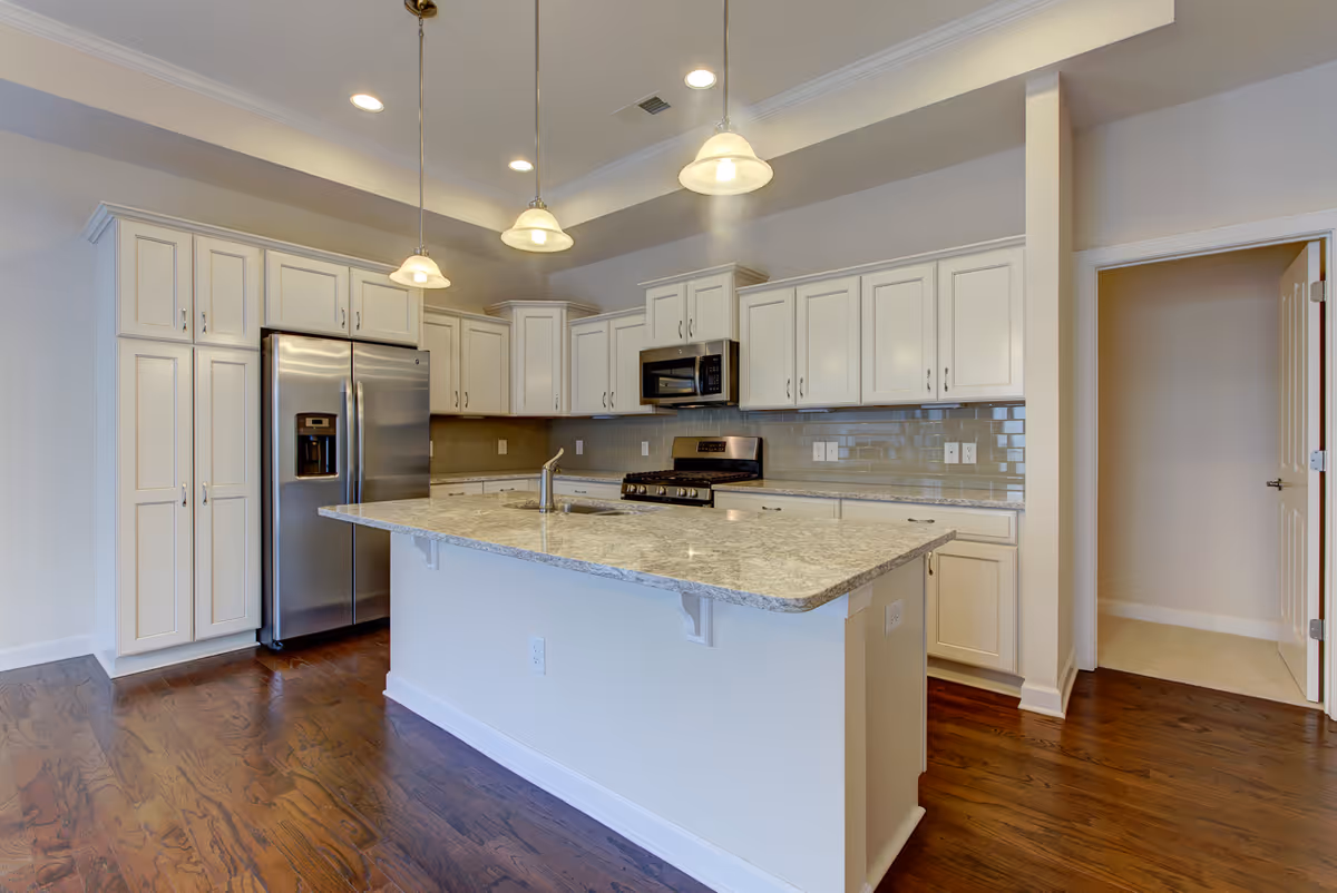 Modern kitchen with white cabinetry, stainless steel refrigerator, stove, and microwave. A large kitchen island with a granite countertop and a sink is centered in the room, illuminated by three hanging pendant lights. The floor is dark hardwood, and there is an open doorway leading to another room.