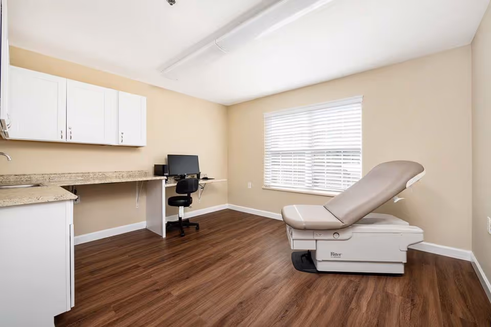 A clean and well-lit medical examination room with beige walls and wood flooring. The room features a beige examination table on the right side, a window with white blinds, white cabinets mounted on the left wall, a long countertop, and a black office chair in front of a computer on a small desk.