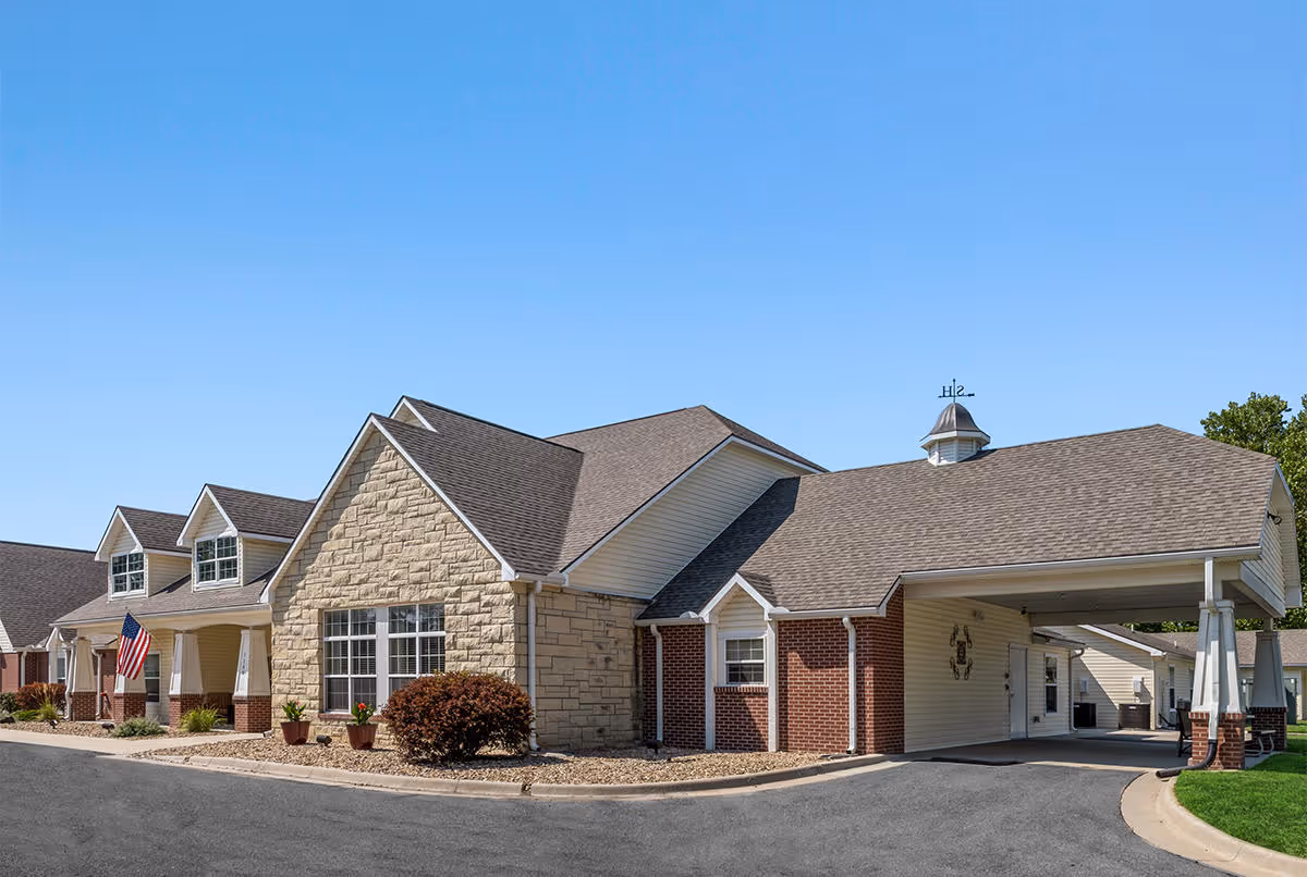 Exterior view of a single-story assisted living facility building with a combination of stone and brick facade, multiple windows, a covered entrance driveway, and an American flag displayed near the front. The sky is clear and blue.