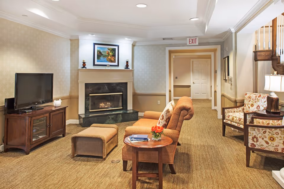 A cozy living room area with a brown upholstered sofa and matching ottoman, a wooden side table with a small vase of red flowers and books, a fireplace with a painting above it, a flat-screen TV on a wooden cabinet, and two floral-patterned armchairs next to a lamp. The room has beige walls, carpeted floor, and a hallway with an exit sign in the background.