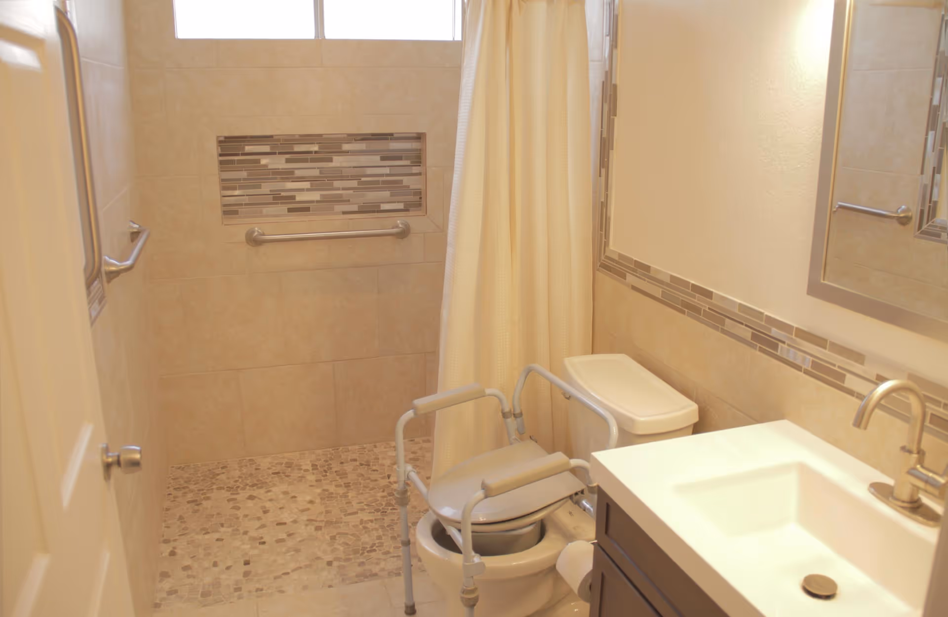 Accessible bathroom with a tiled walk-in shower, grab bars, shower curtain, raised commode chair over the toilet, and a vanity sink.