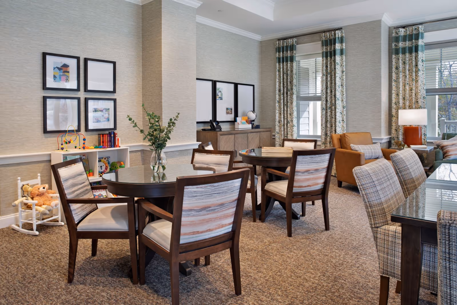 A cozy common area in a senior living facility featuring two round tables with wooden chairs, a small white bookshelf with children's toys and books, framed artwork on the wall, patterned curtains on windows, a brown armchair, and a lamp on a side table.