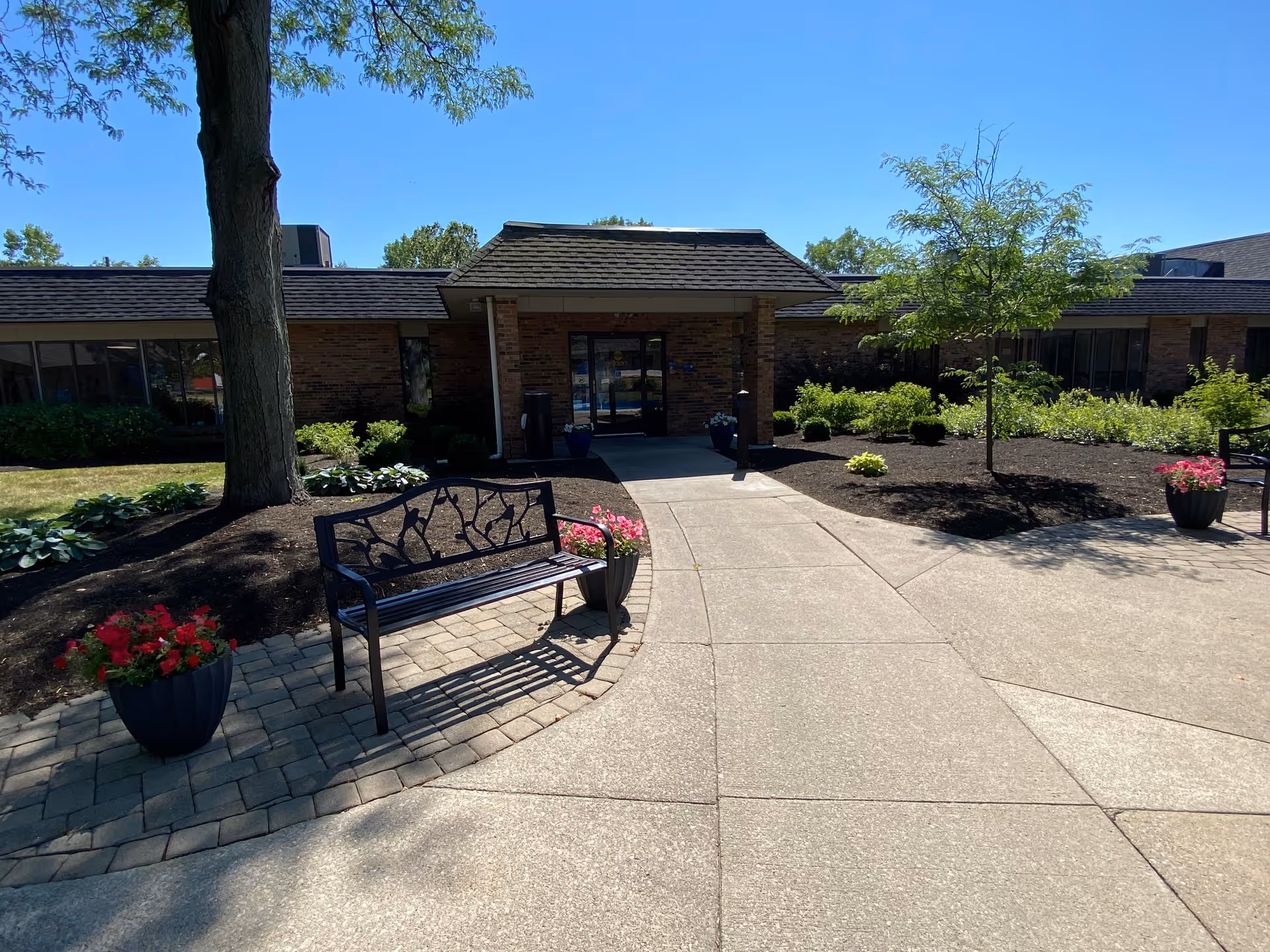 Outdoor view of the entrance to Brickyard Healthcare - Muncie Care Center on a sunny day. The entrance features a covered porch with glass doors, surrounded by landscaped areas with trees, shrubs, and flower pots. A decorative metal bench is placed on a paved area near the walkway leading to the entrance.