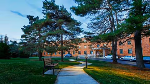 Brick senior living facility exterior with trees, a walkway, benches, and parked cars.