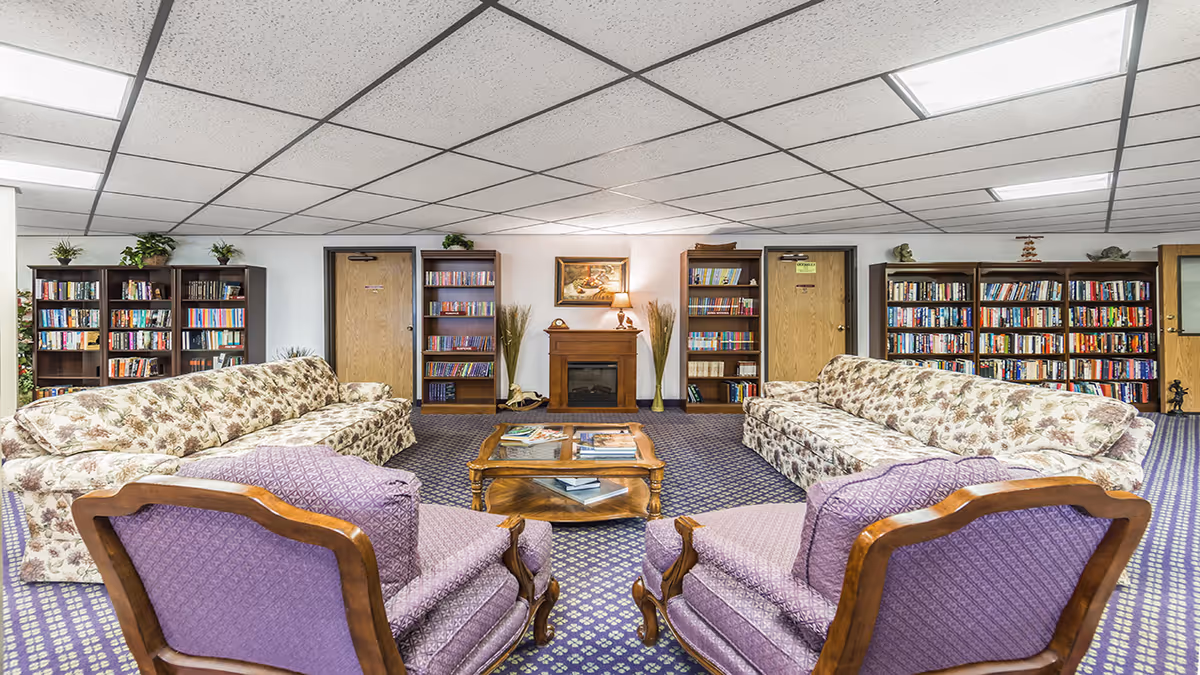 A cozy common area with two floral-patterned sofas facing each other, two purple upholstered armchairs, a wooden coffee table with books, multiple bookshelves filled with books, a small fireplace with a lamp and painting above it, and a carpeted floor with a patterned design.