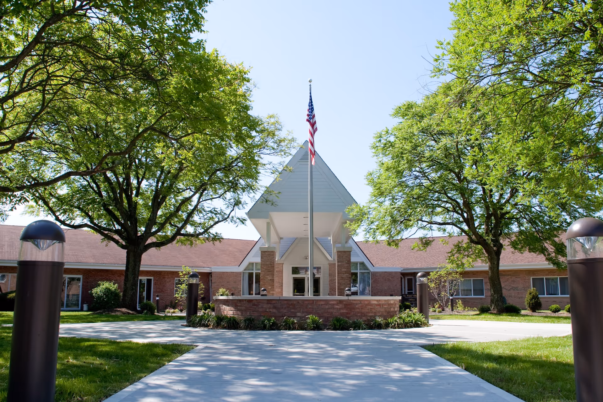 Front exterior view of St. Paul Village facility with a paved walkway leading to the entrance, an American flag on a flagpole in the center, surrounded by green trees and lawn under a clear blue sky.