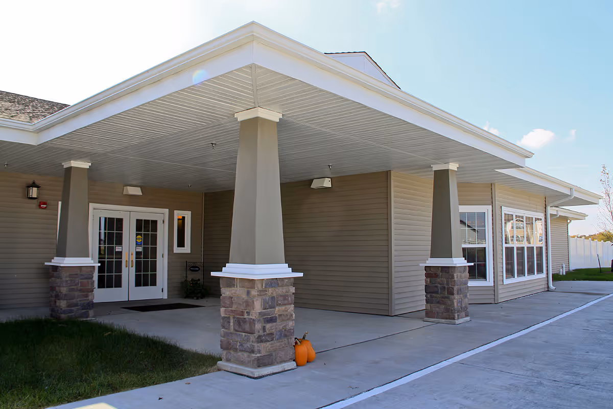 Covered front entrance of a single-story building with columns, double glass doors, and pumpkins by the curb.