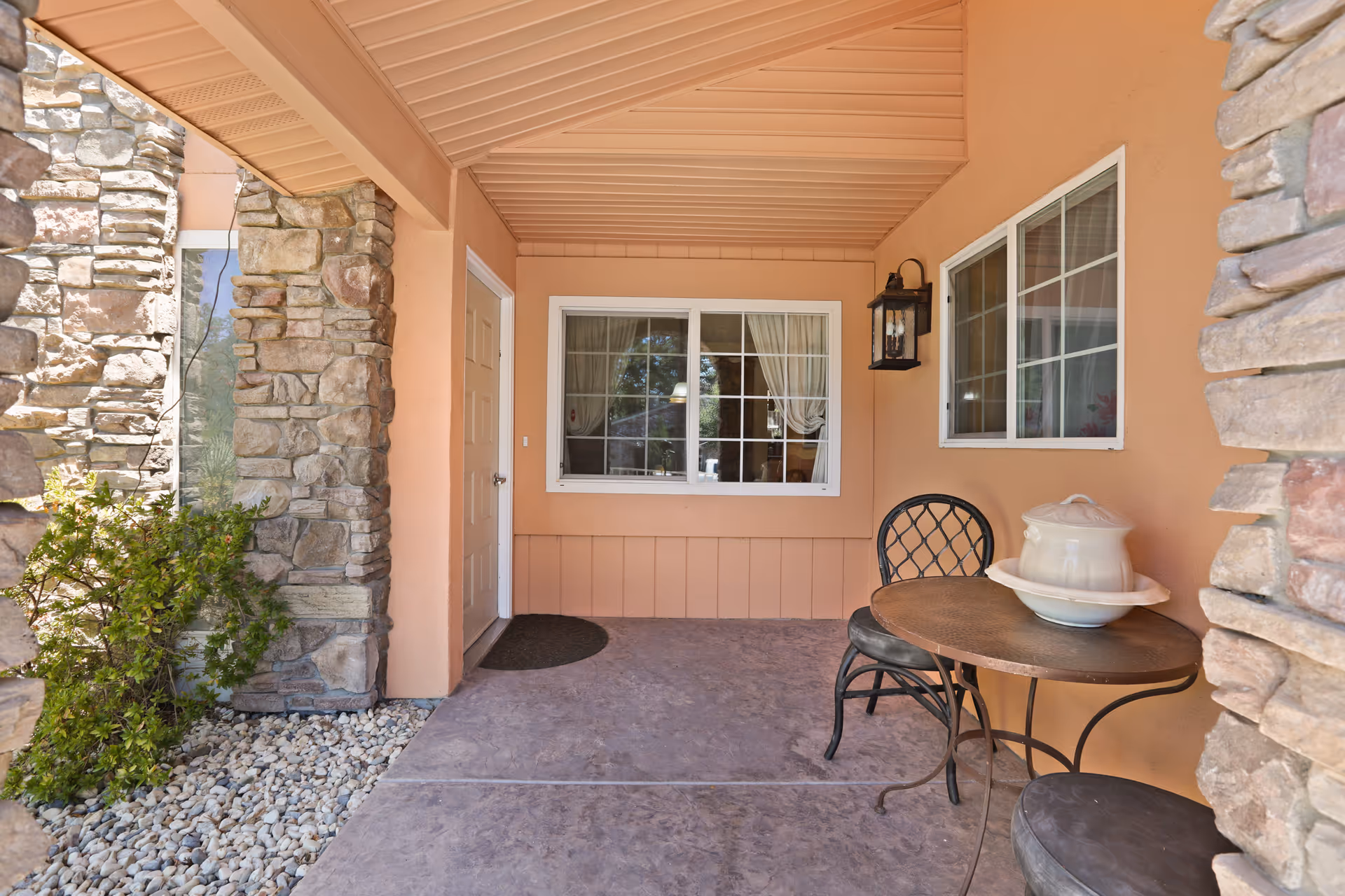 Covered front porch with stone columns, a small metal table and chairs, and windows on a peach-colored building.