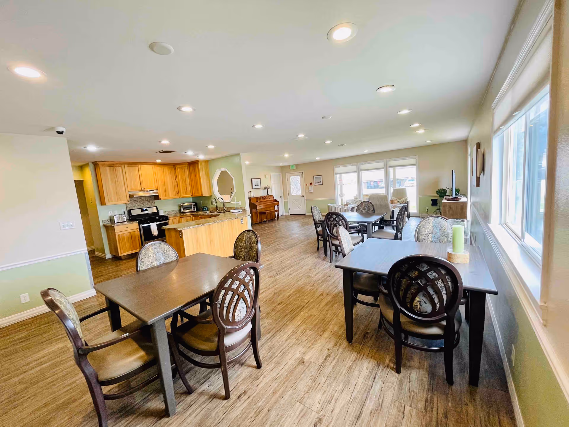 A spacious, well-lit common area in a senior living facility featuring a kitchen with wooden cabinets and stainless steel appliances on the left, multiple dining tables with chairs arranged throughout the room, large windows on the right letting in natural light, and a piano against the far wall near the entrance door.