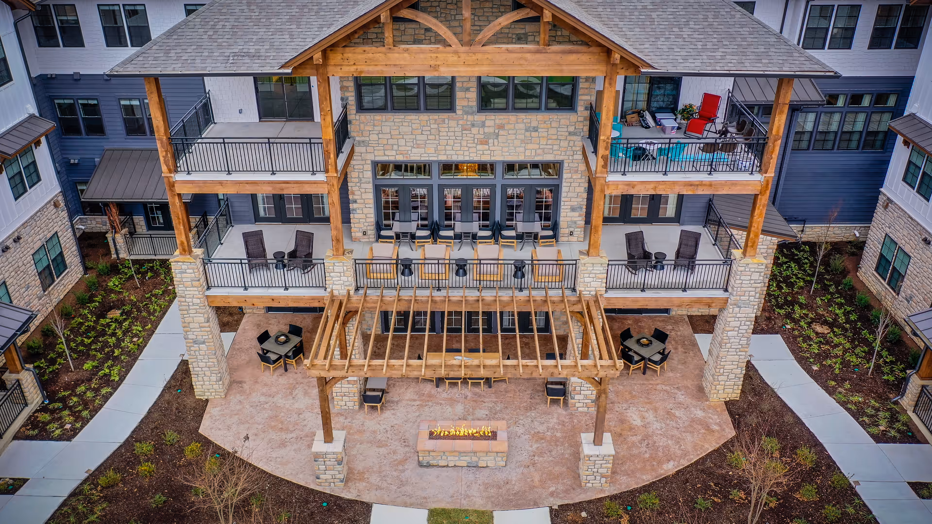 Outdoor patio area of a senior living facility featuring a large wooden pergola with seating underneath, a rectangular fire pit, multiple tables with chairs, and balconies with additional seating on the upper floors. The building exterior is a combination of stone and siding with large windows, surrounded by landscaped walkways and greenery.
