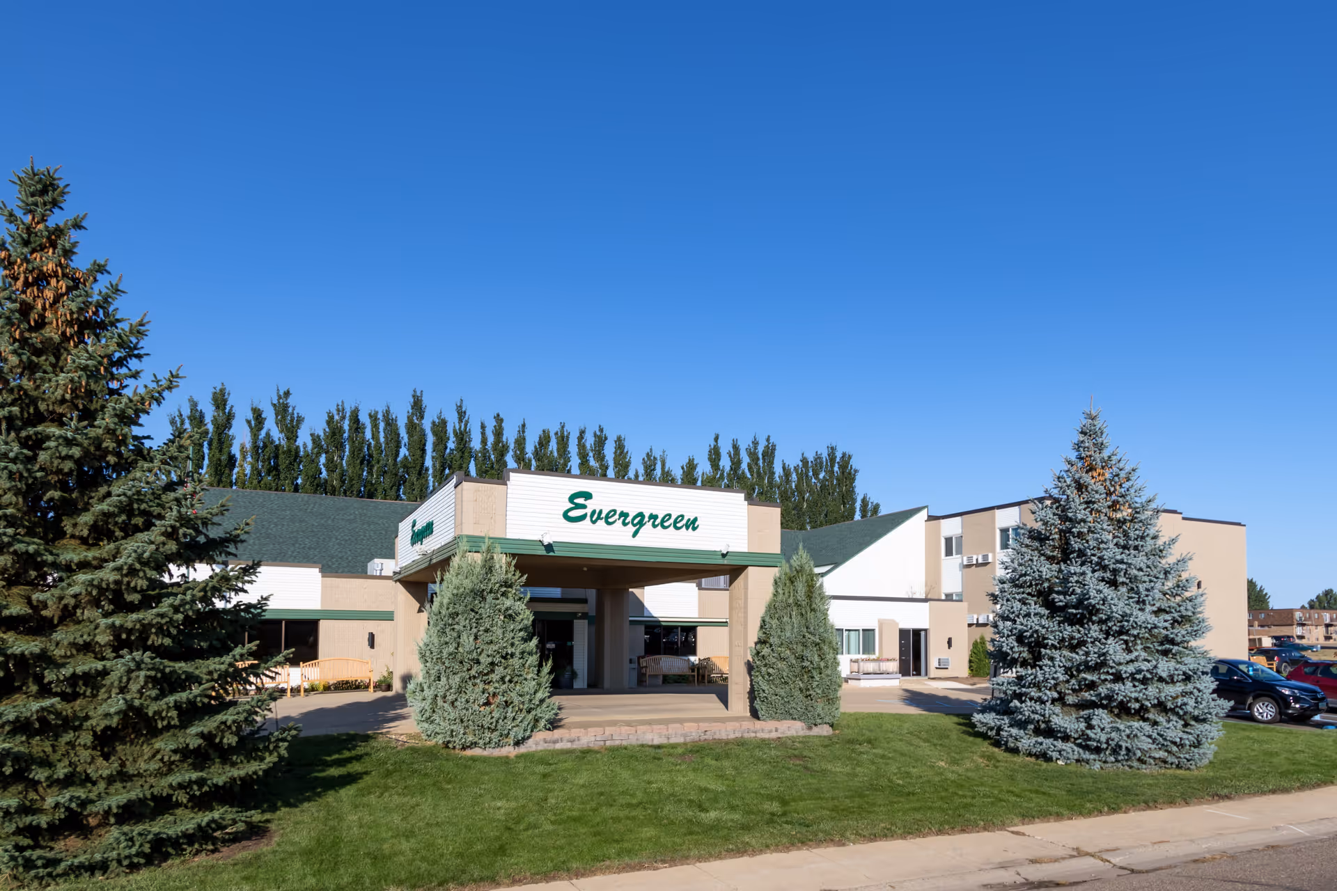 Exterior view of the Evergreen senior living facility building with a clear blue sky, green lawn, and several evergreen trees in front.