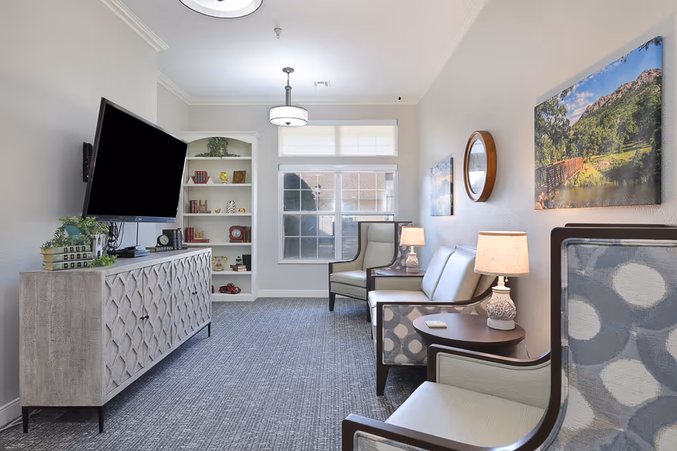 A bright and neatly arranged living room with a mounted flat-screen TV on the left wall above a decorative wooden cabinet. The room features a carpeted floor, a window letting in natural light, a white built-in bookshelf with decorative items, and several upholstered chairs and a loveseat along the right wall. Two table lamps and framed landscape artwork decorate the space.