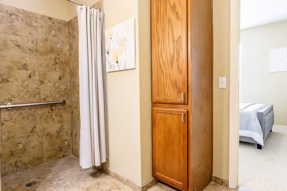 A bathroom with a tiled walk-in shower featuring a grab bar and a white shower curtain. Adjacent to the shower is a wooden cabinet with two doors. In the background, a bedroom with a bed and light-colored walls is partially visible through an open doorway.