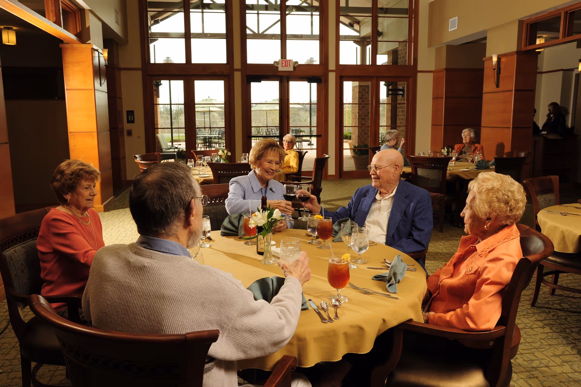 Several elderly residents sit around a round dining table in a bright dining room, smiling and toasting with drinks.