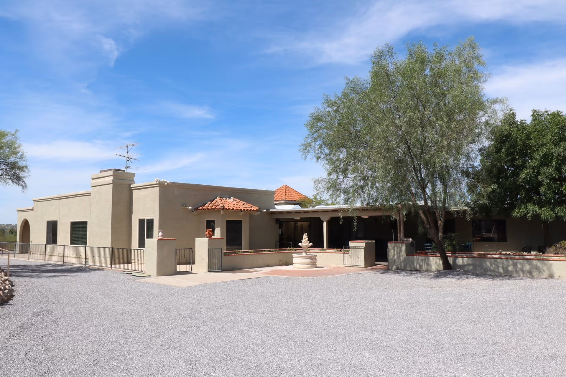 Single-story stucco building with a courtyard fountain, gravel driveway and trees under a blue sky.