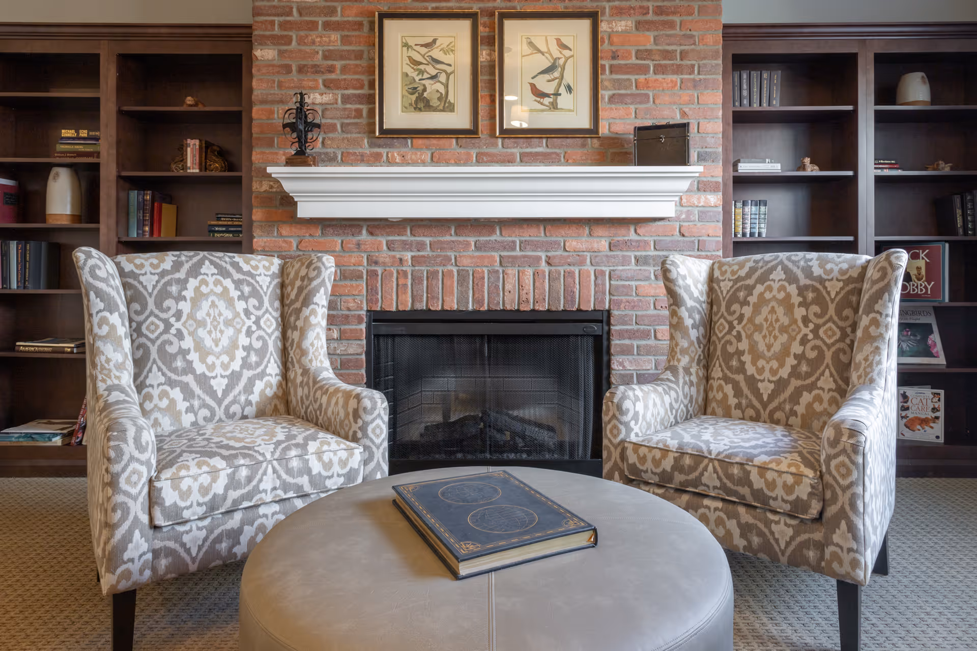 Two patterned armchairs facing a round ottoman with a closed book on it, positioned in front of a brick fireplace with a white mantel. Above the mantel are two framed bird illustrations. On either side of the fireplace are built-in dark wood bookshelves with books and decorative items.