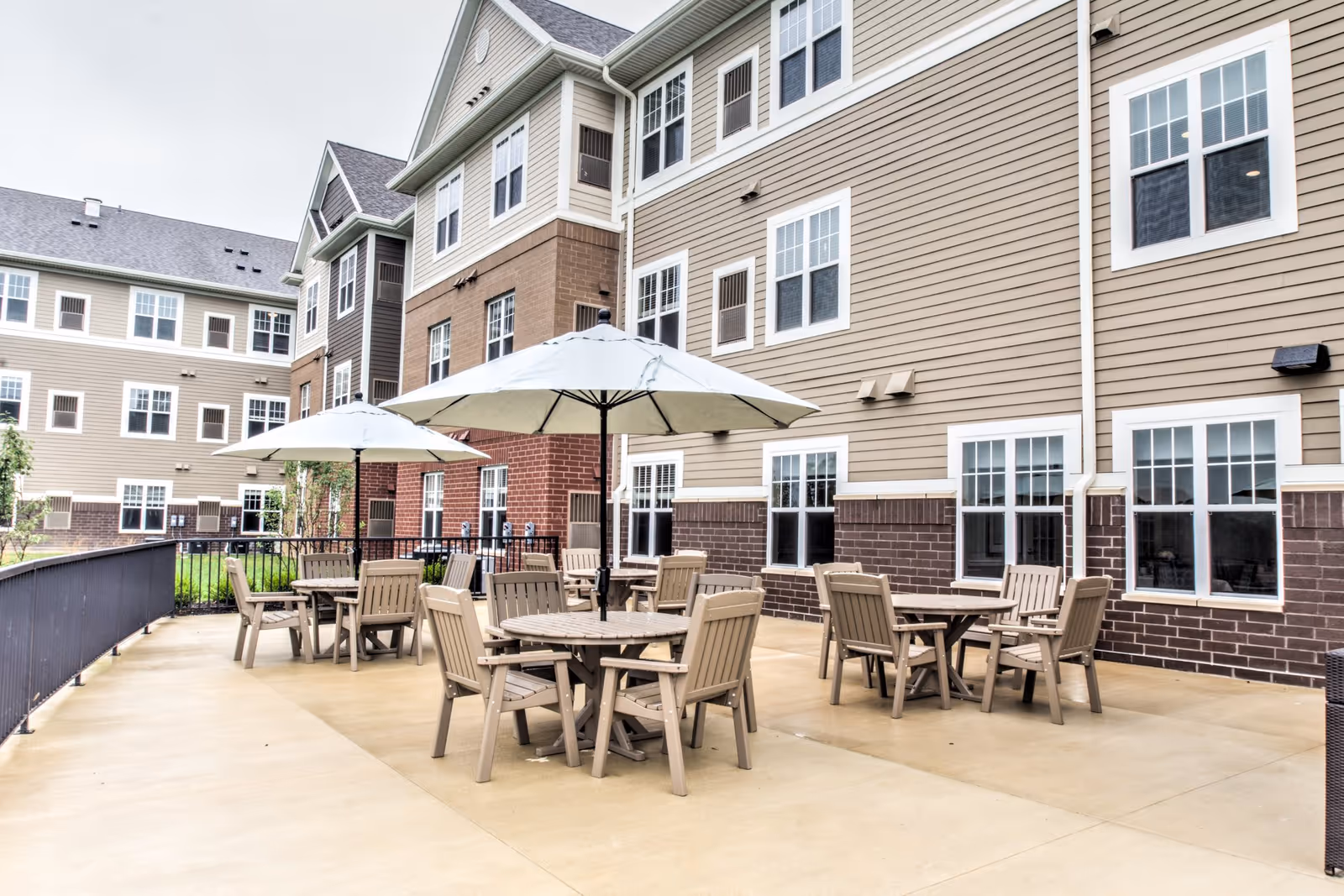Outdoor patio area with round tables, chairs, and umbrellas beside a multi-story senior living building.