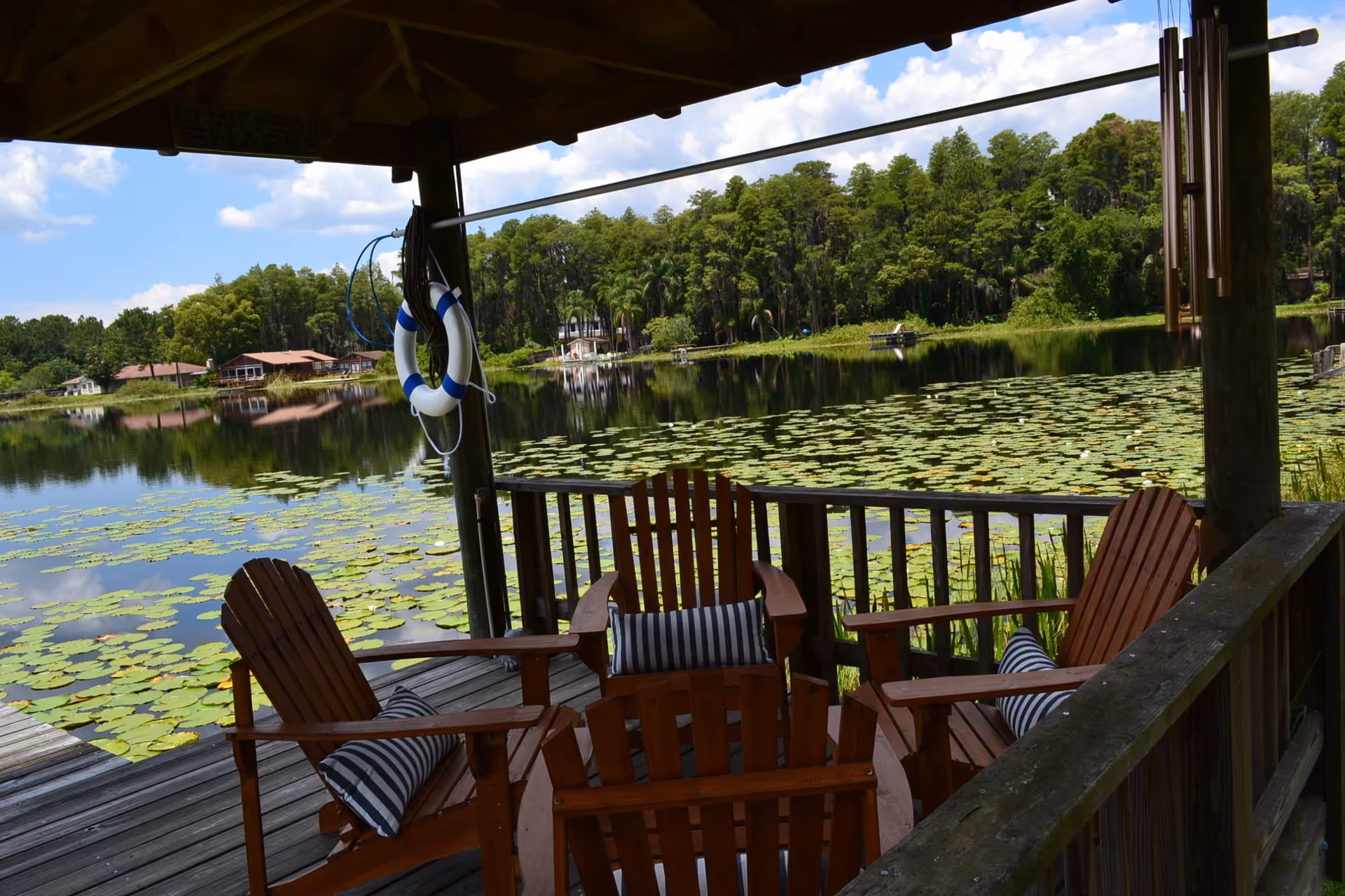 A wooden dock with four Adirondack chairs and striped cushions under a shaded roof overlooking a lake covered with lily pads. A life preserver hangs on a post, and there are trees and houses visible across the water under a partly cloudy sky.