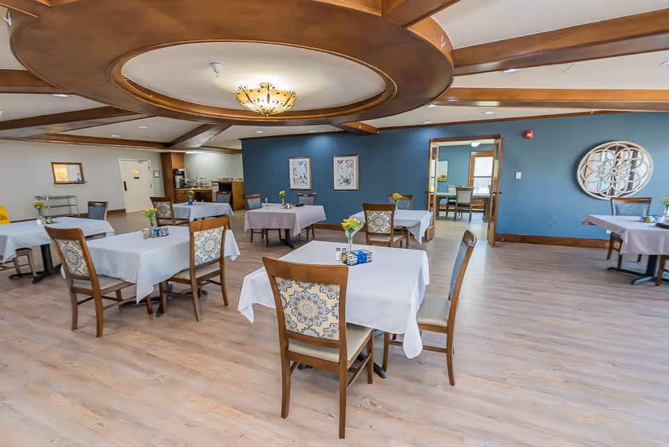 A spacious dining room with several tables covered in white tablecloths, each surrounded by wooden chairs with patterned cushions. The room features a wooden ceiling design with a central light fixture, light wood flooring, and blue accent walls decorated with framed artwork and a decorative mirror. There is an open doorway leading to another room with additional seating.
