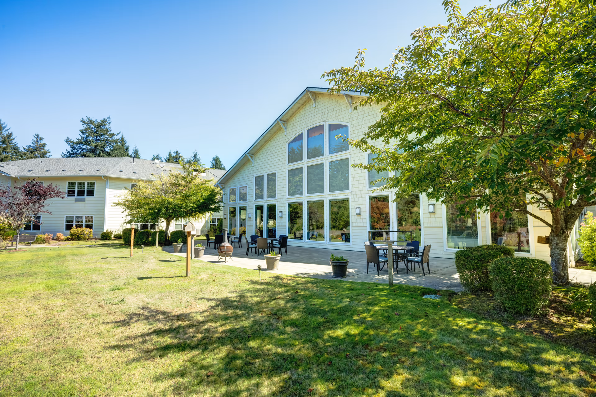 Exterior view of a senior living facility building with large windows and a patio area furnished with tables and chairs. The building is surrounded by a well-maintained lawn, trees, and shrubs under a clear blue sky.
