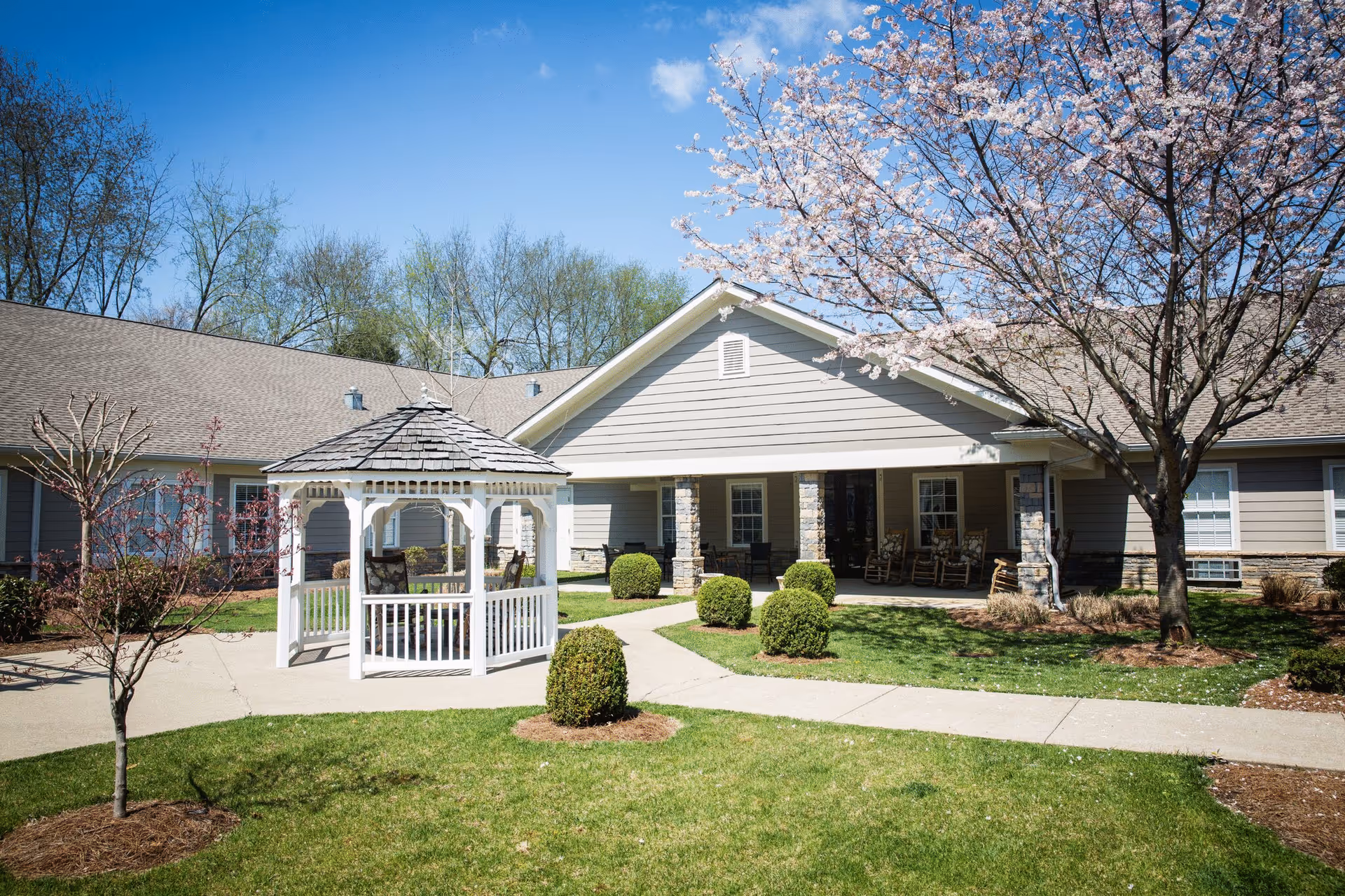 Outdoor courtyard area of Chandler Park Assisted Living featuring a white gazebo with chairs inside, a paved walkway, manicured bushes, a blooming tree, and a building with a covered porch and rocking chairs under a clear blue sky.