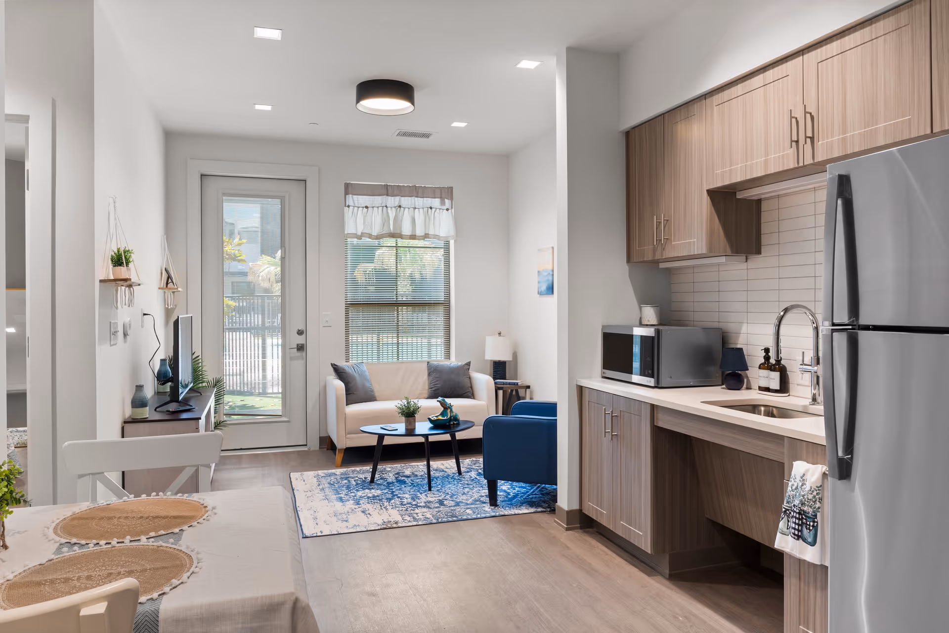 A modern, open-concept living space featuring a small kitchen area with wooden cabinets, a stainless steel refrigerator, a microwave, and a sink. Adjacent to the kitchen is a cozy living room with a white loveseat, a blue armchair, a small coffee table on a blue patterned rug, and a TV on a wooden stand. A door and window with blinds provide natural light to the room.