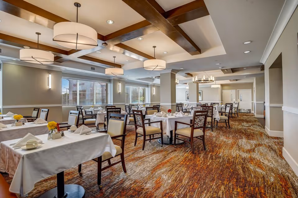 A spacious dining room with multiple tables covered in white tablecloths, each set with napkins, cups, and small flower arrangements. The room features large windows with blinds, modern ceiling lights, and wooden beams on the ceiling. The carpet has a multicolored pattern in shades of brown, orange, and beige.