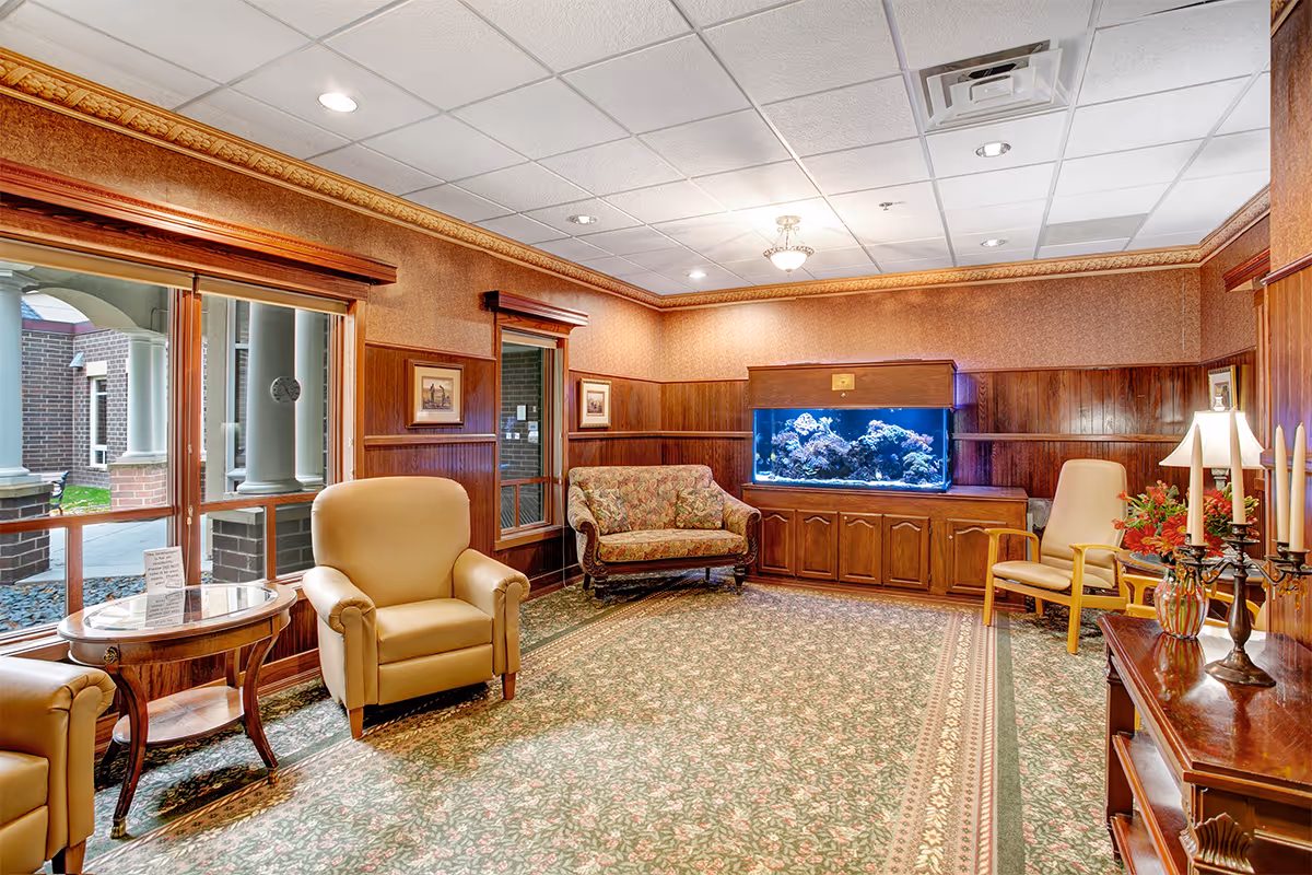 A cozy sitting area inside Boutwells Landing featuring beige armchairs, a floral upholstered loveseat, a large aquarium built into a wooden cabinet, a wooden side table with a glass top, and a decorative lamp with candles. The room has patterned carpet, wood-paneled walls, framed artwork, and a window looking out to a covered outdoor area with brick columns.