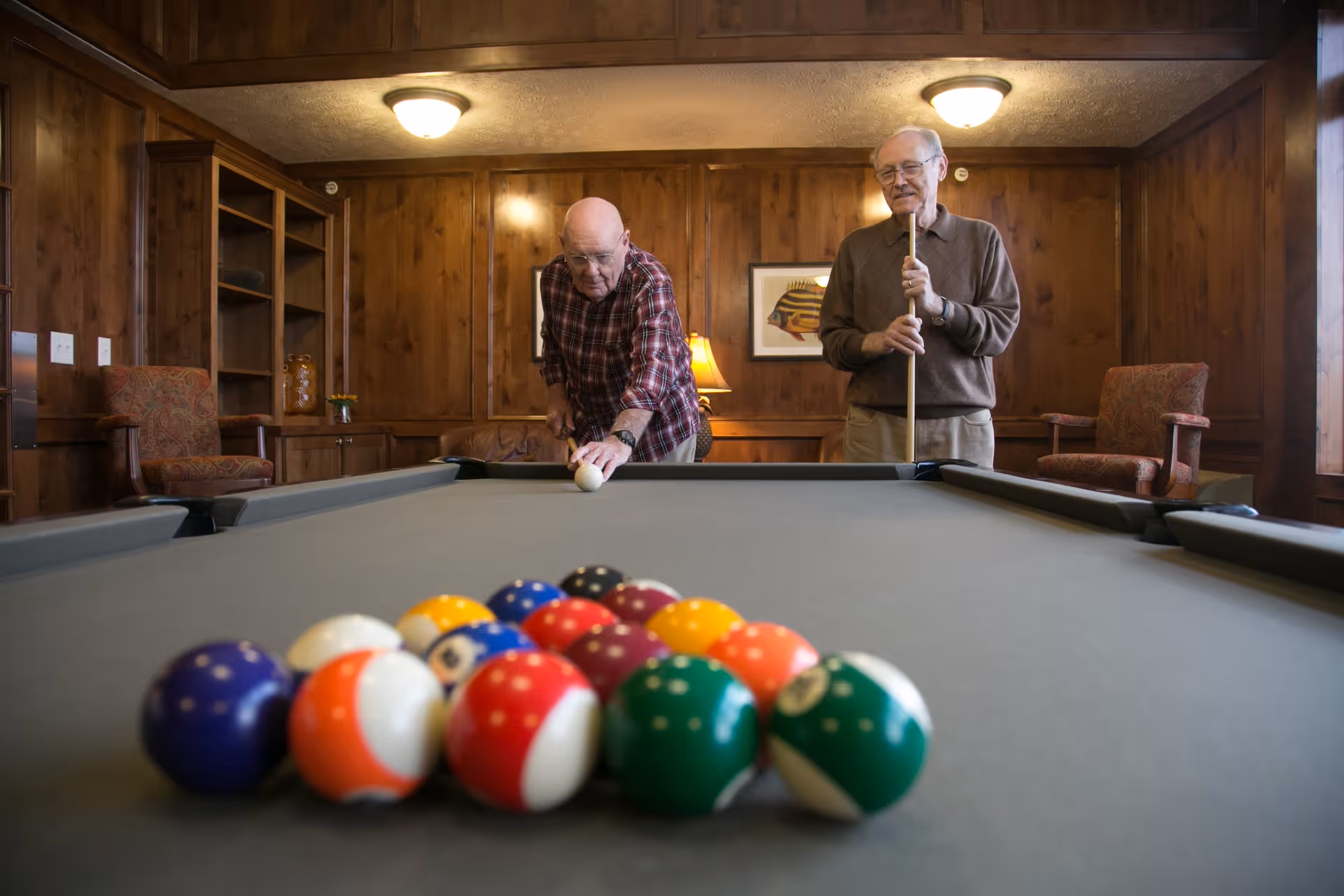 Two elderly men playing pool in a wood-paneled room with comfortable chairs and framed artwork on the walls. One man is preparing to take a shot while the other watches, holding a pool cue.