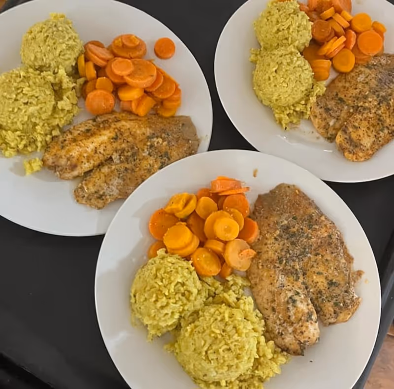 Three white plates each containing two scoops of yellow rice, cooked sliced carrots, and seasoned grilled chicken breast on a black surface.