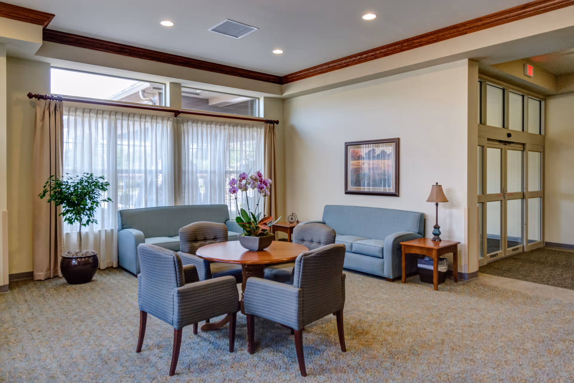 A bright and spacious common area with two blue sofas, a round wooden table surrounded by four upholstered chairs, a potted plant, a table lamp on a side table, and large windows with sheer curtains allowing natural light to fill the room.