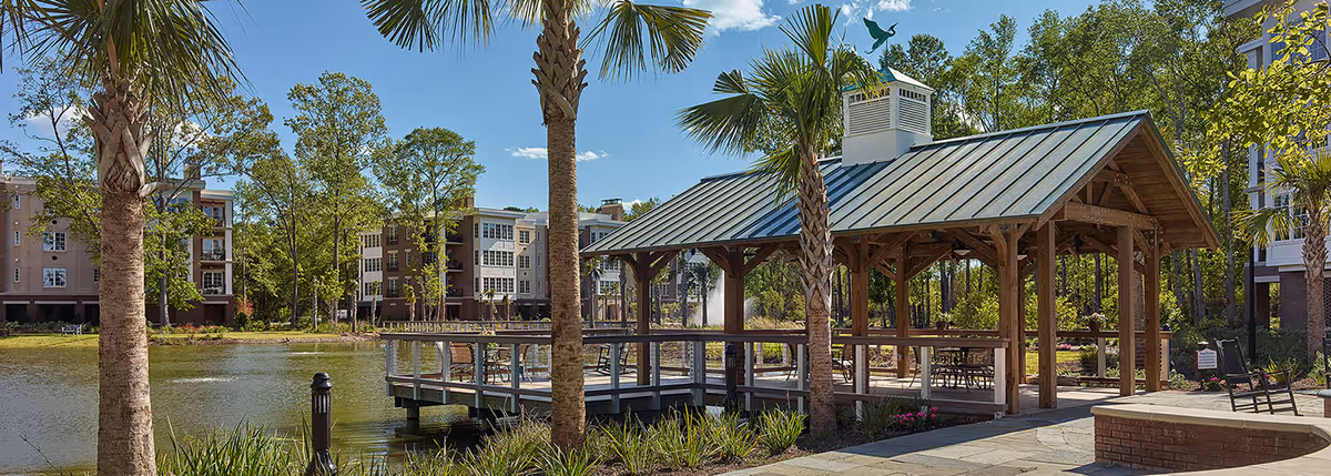 A wooden lakeside pavilion with a green metal roof on a dock, flanked by palm trees and apartment buildings across the water.