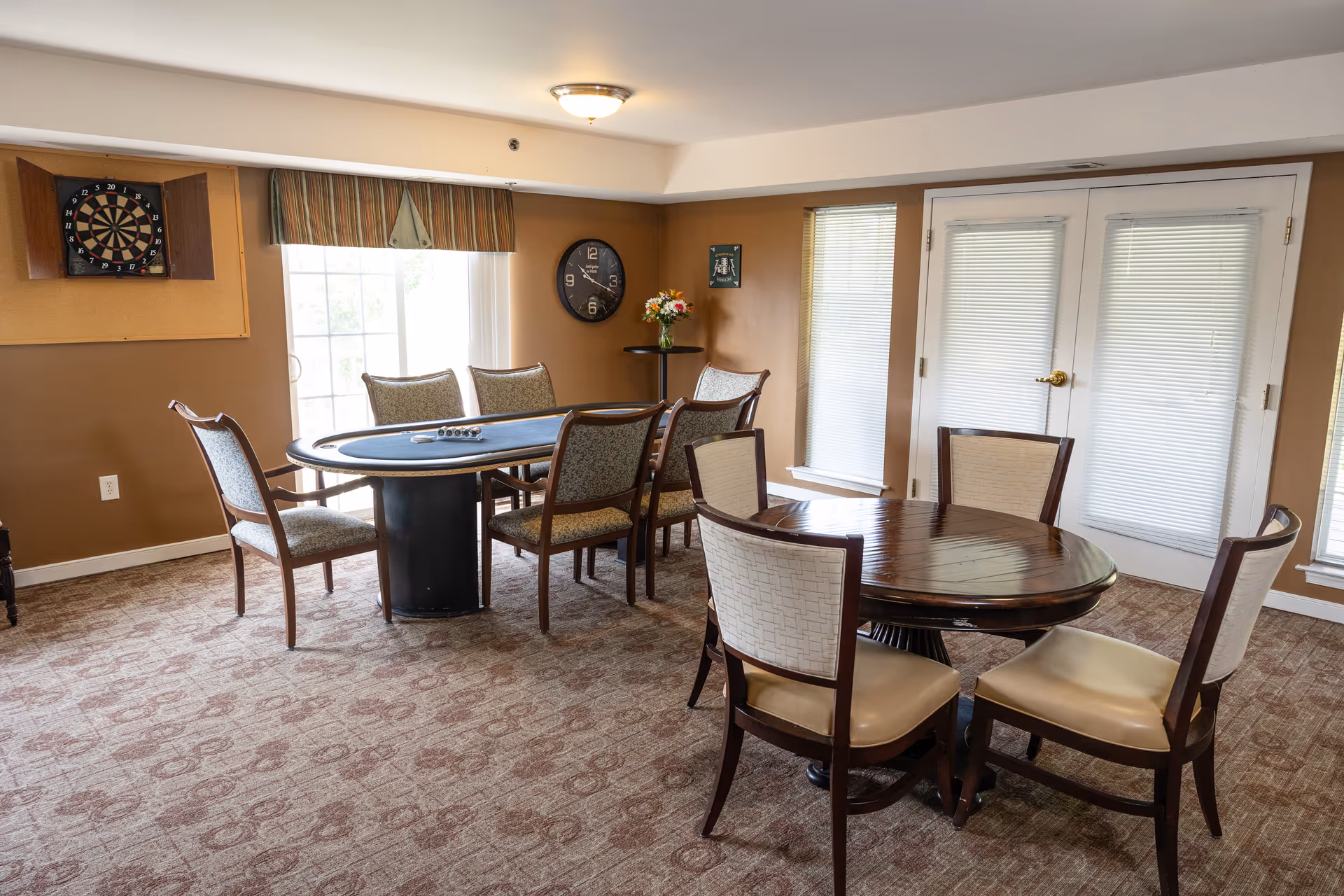 A well-lit room with two tables and several chairs arranged around them. One table is oval with a blue felt surface, likely for card games, surrounded by six wooden chairs with patterned cushions. The other table is round with a polished wooden surface and four chairs with cream-colored cushions. The room has beige walls, a dartboard mounted on the wall, a clock, a small round table with a vase of flowers, and large windows with blinds and a glass door letting in natural light.