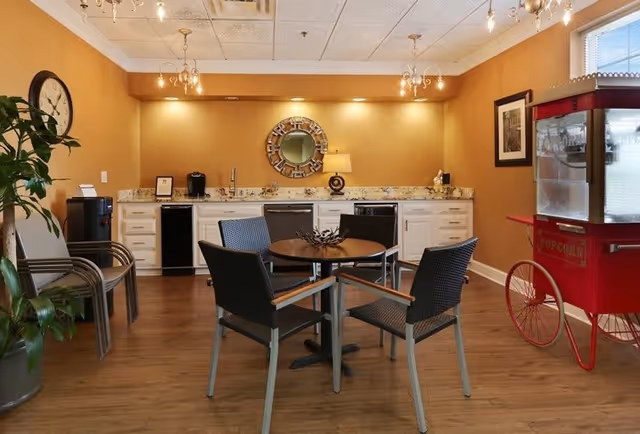 A cozy interior room with a round table surrounded by four chairs in the center. The back wall features a countertop with cabinets, a coffee maker, a lamp, and a decorative round mirror. To the right, there is a vintage-style red popcorn machine. The room has warm beige walls, wooden flooring, and ceiling lights.