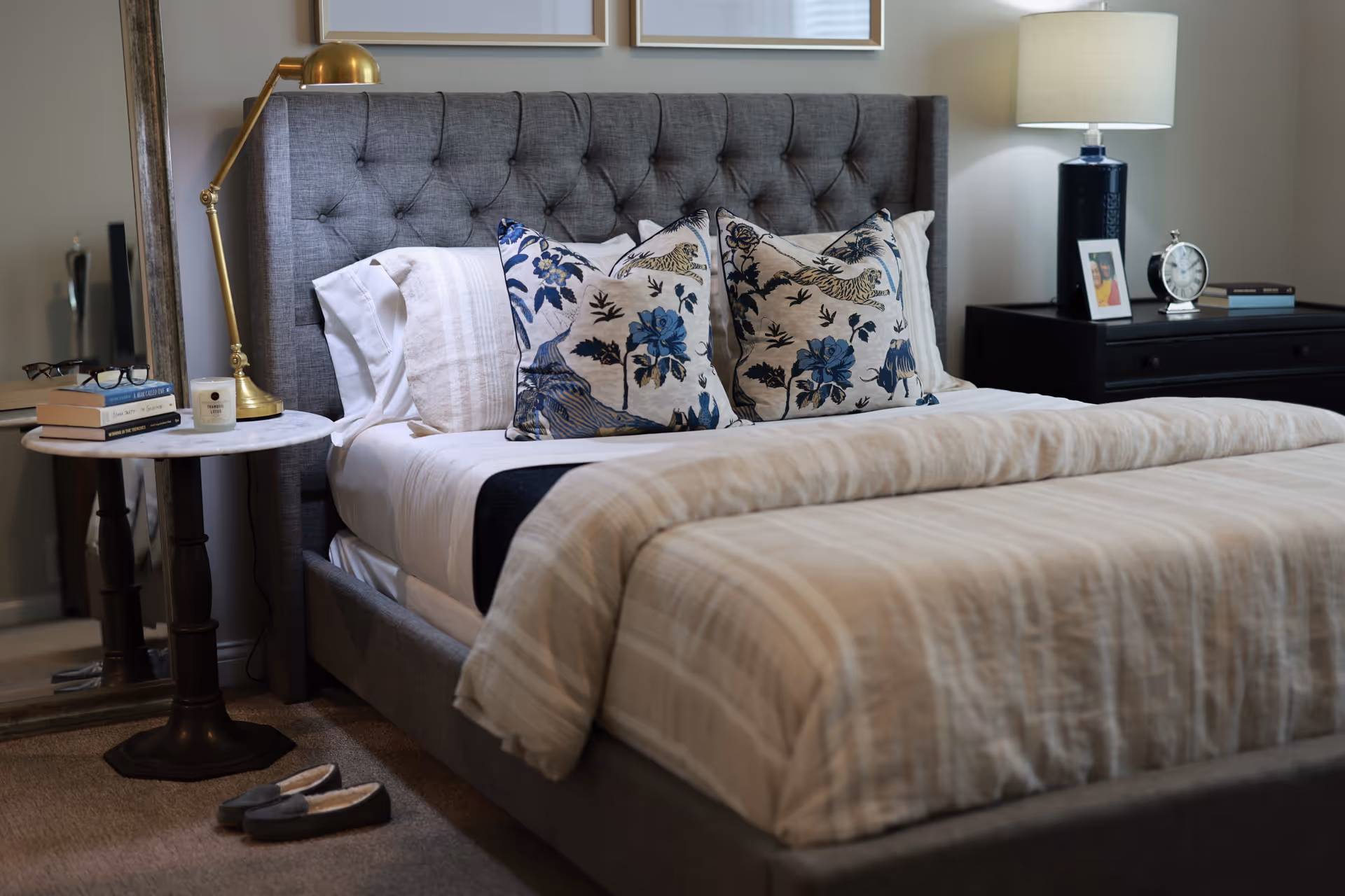 Neatly made bedroom with a tufted gray headboard, patterned throw pillows, bedside tables, lamps, and slippers on the floor.