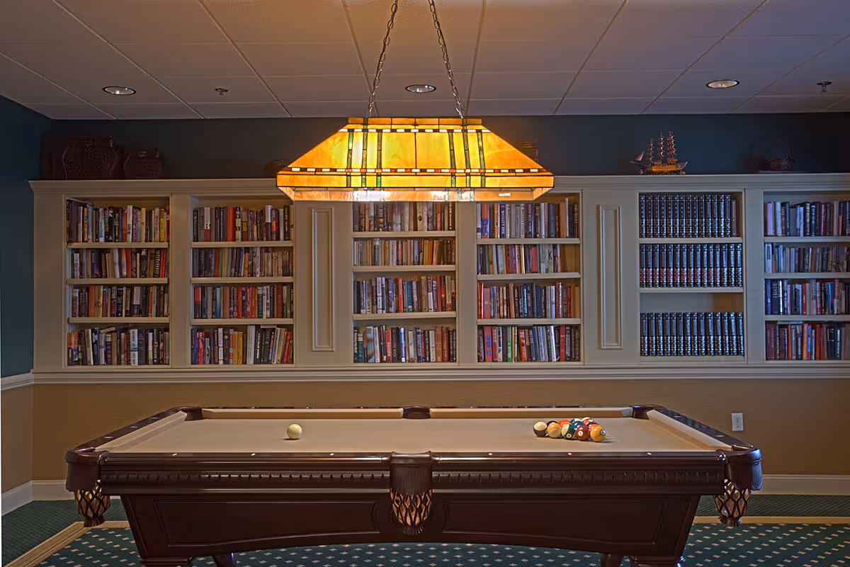 A cozy indoor room featuring a pool table with billiard balls arranged on it, positioned in front of a large built-in bookshelf filled with numerous books. Above the pool table hangs a rectangular stained glass pendant light, casting a warm glow. The room has a carpeted floor with a patterned design and a ceiling with recessed lighting.