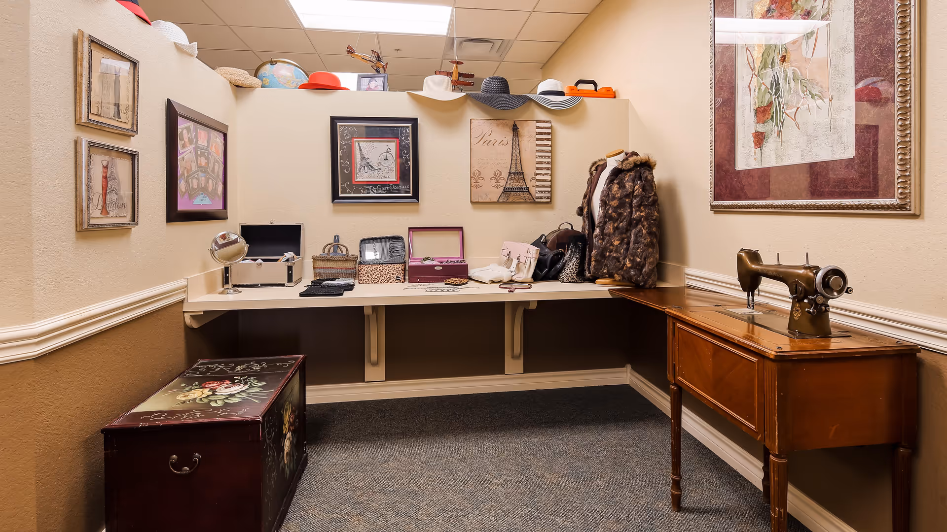 A cozy sewing and craft corner in a senior living facility featuring a vintage sewing machine on a wooden table, a wall-mounted shelf with various hats displayed on top, framed artwork including images of the Eiffel Tower, and a decorative chest with floral designs on the floor.