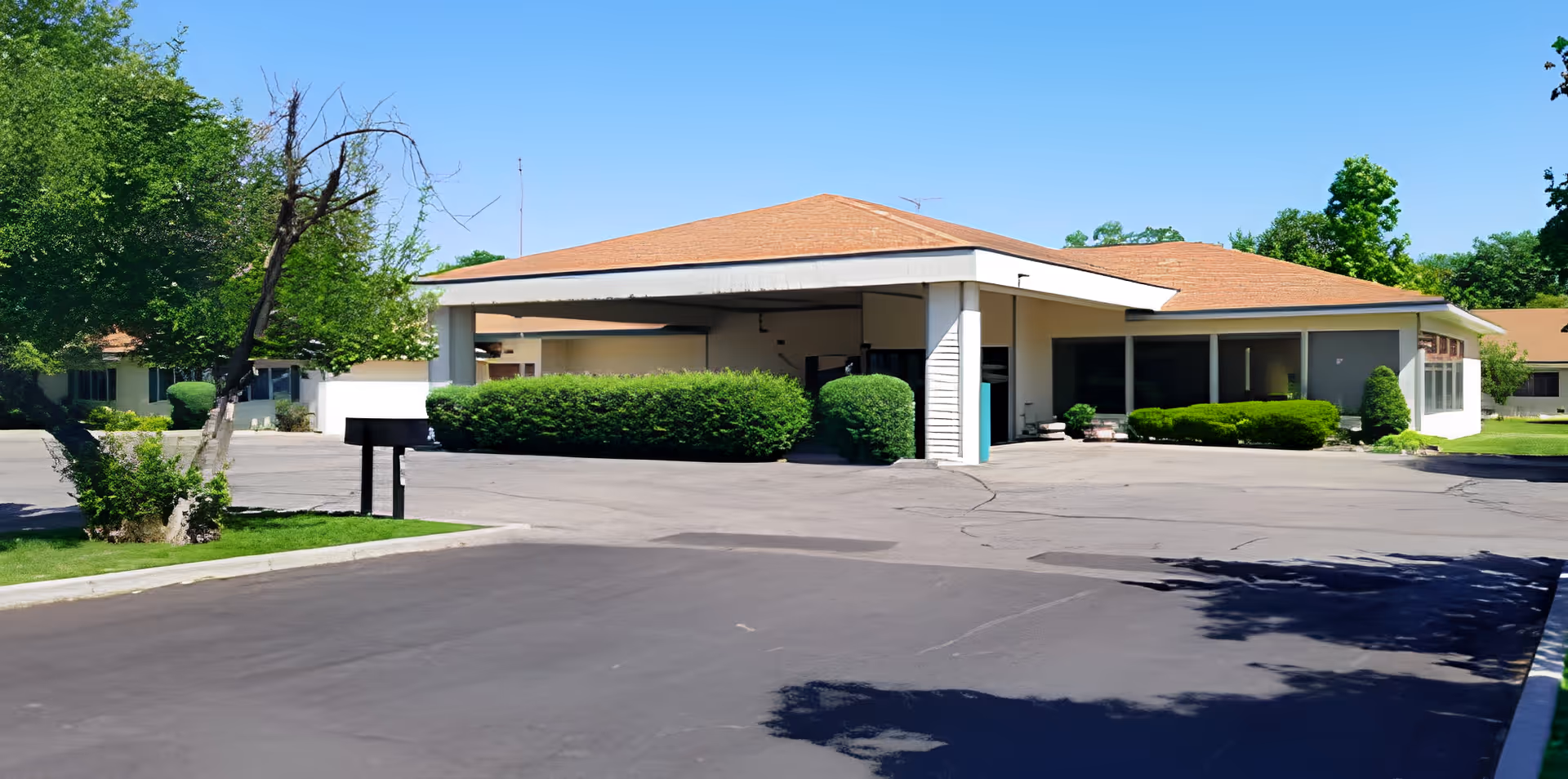 Exterior view of a single-story assisted living or retirement home building with a covered entrance, surrounded by green bushes and trees under a clear blue sky.