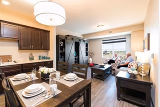 A cozy living and dining area in a senior living facility. The dining table is set with plates, bowls, glasses, and silverware for four people. The living room features a sofa with two elderly people sitting and looking out the window. There is a coffee table, a dark wood entertainment center, and a side table with a lamp. The room has wooden flooring and neutral-colored walls with a large window letting in natural light.