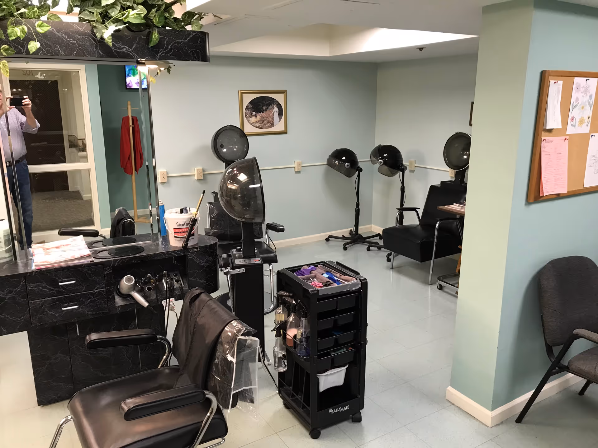 Interior view of a hair salon area in a senior living facility with black salon chairs, hair dryers, a rolling cart with hair styling tools, a large mirror, and a bulletin board on a light blue wall.